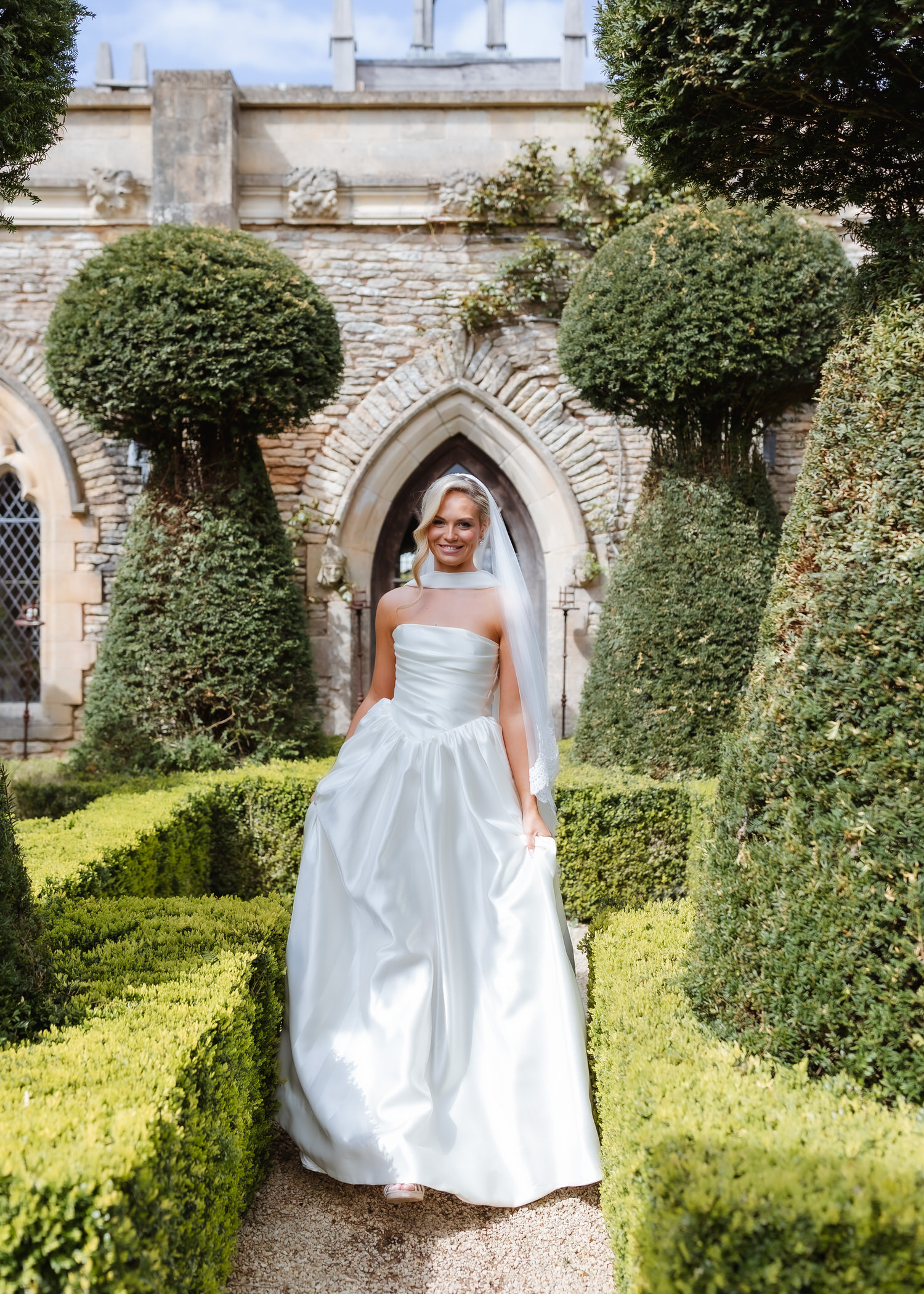 A bride in a white wedding dress and veil smiles while walking through a formal garden, with tall, sculpted hedges and an old stone building—perfect for a country-house wedding photographer to capture timeless moments.