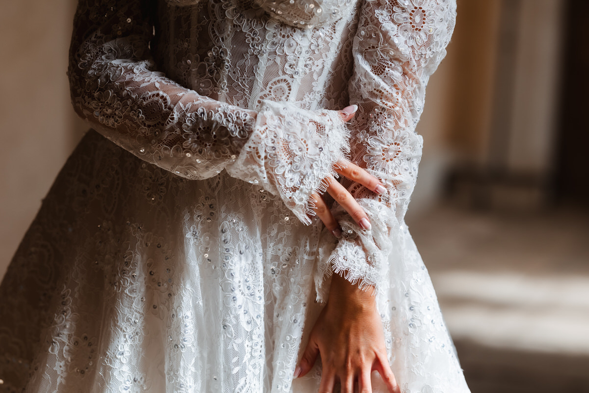A close-up of a person wearing a detailed, long-sleeved white lace wedding dress, gently touching their own arm, with soft natural light highlighting the intricate patterns—captured beautifully by a Hampshire wedding photographer.