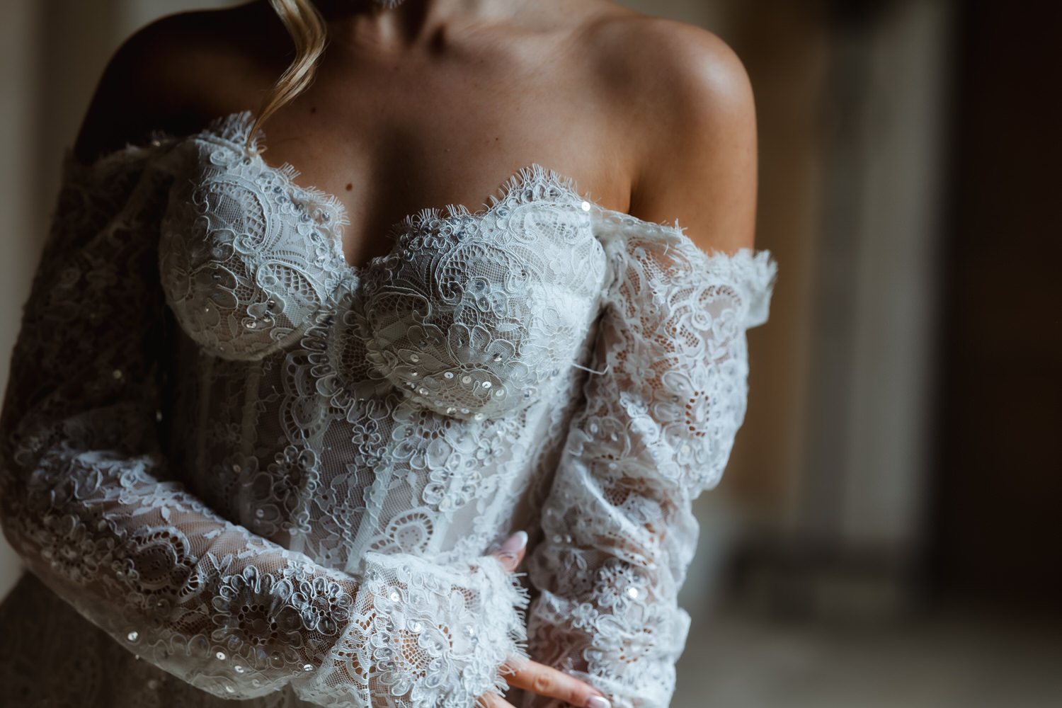 A woman in an off-the-shoulder, lace wedding dress with intricate embroidery and beadwork poses gracefully. Captured by a country-house wedding photographer, soft lighting enhances the gown’s delicate texture as she crosses her arms at her waist.