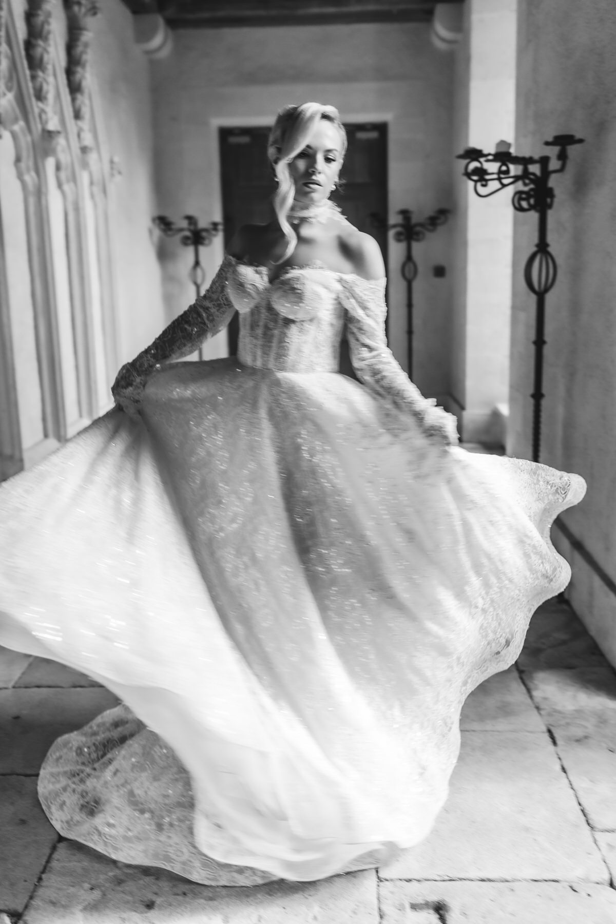 Black and white photo of a woman in an elegant, off-the-shoulder gown twirling indoors, her dress billowing as she moves gracefully on a stone floor with arched columns—a timeless shot by a Hampshire wedding photographer.