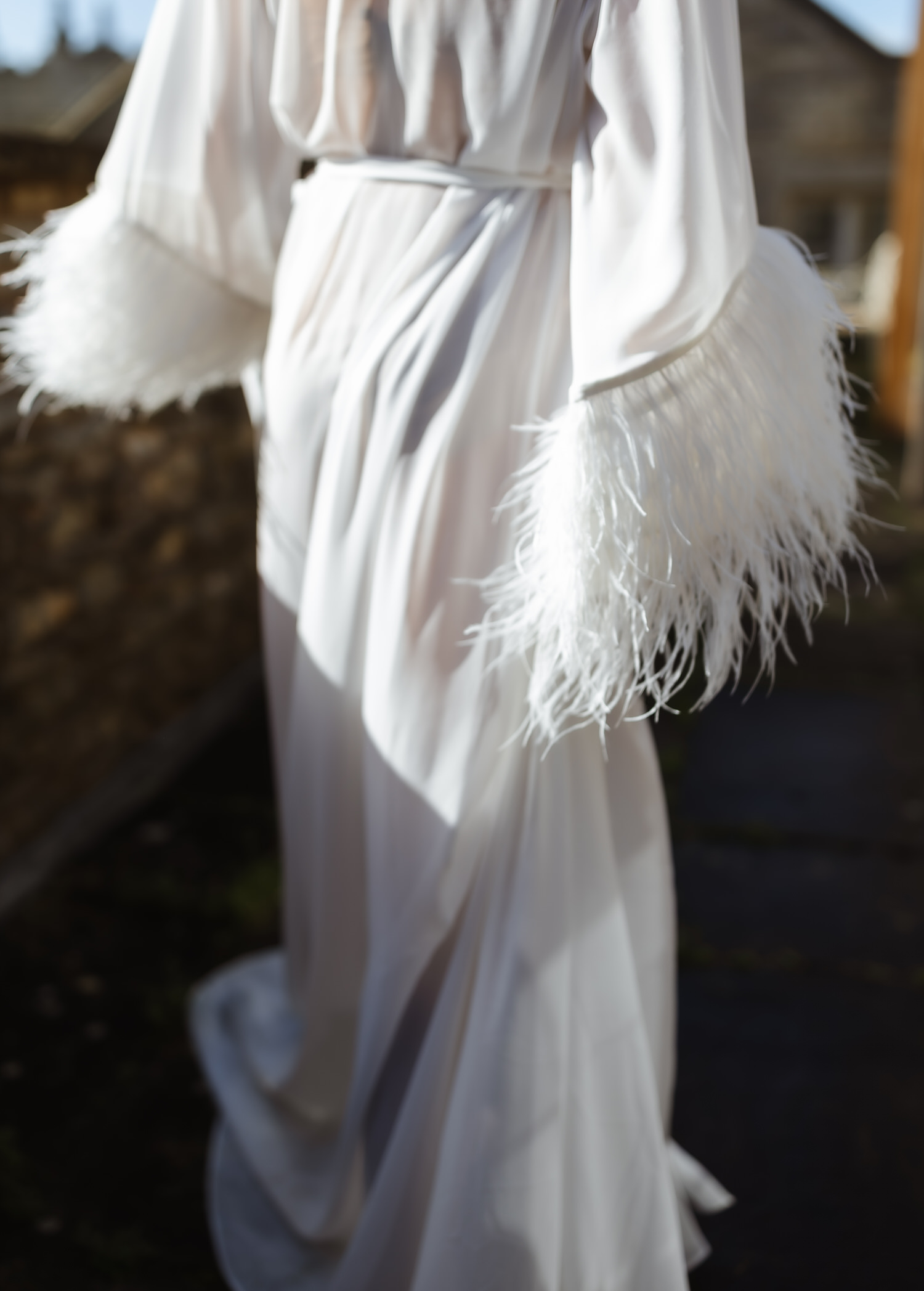 A person wearing a long, flowing white robe with feathered cuffs walks outdoors on a sunny day, the softly blurred background evoking the dreamlike style of a country-house wedding photographer.