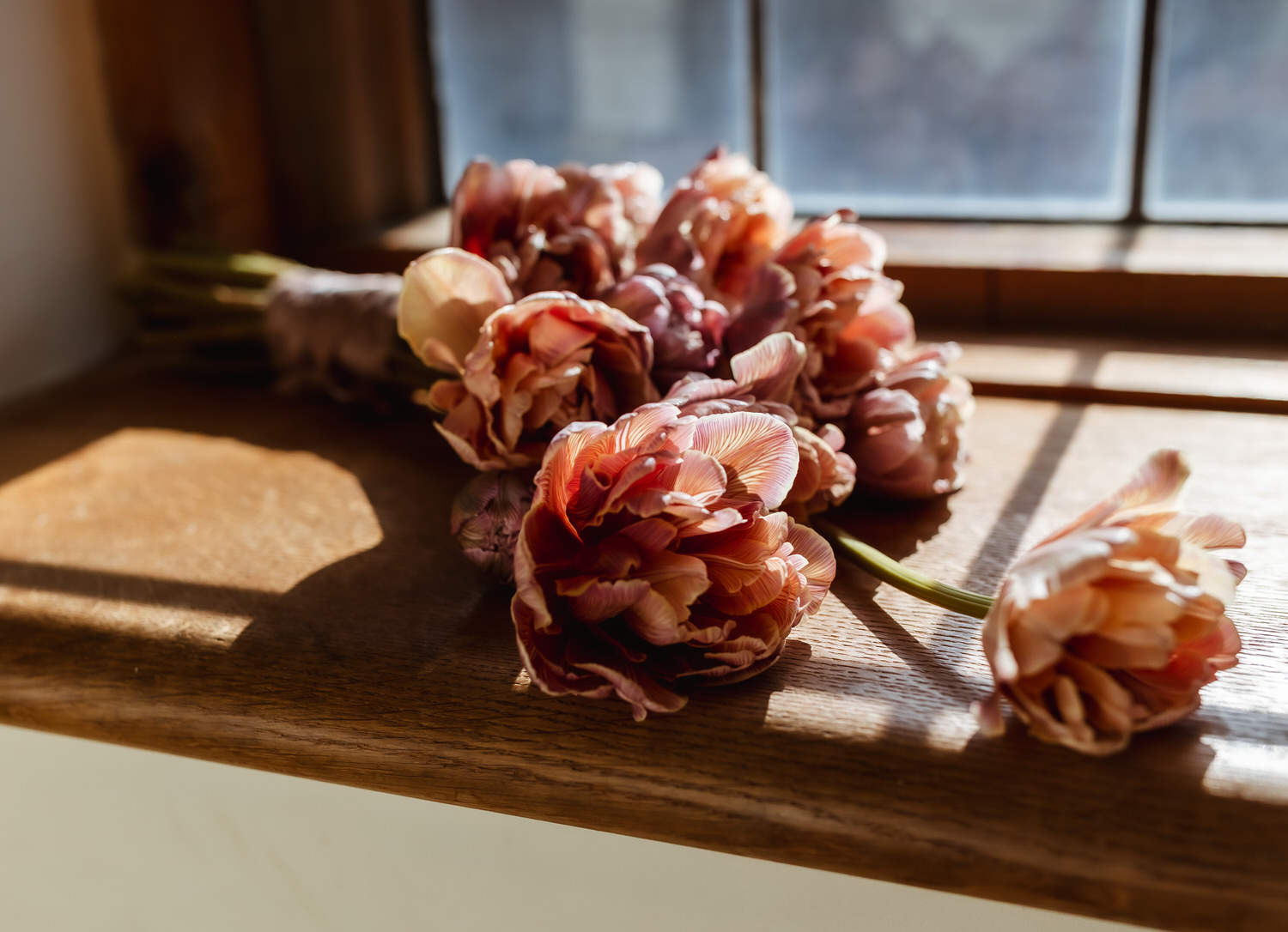 A bouquet of pinkish-orange tulips lies on a wooden windowsill, illuminated by soft sunlight—a scene reminiscent of moments captured by a country-house wedding photographer.