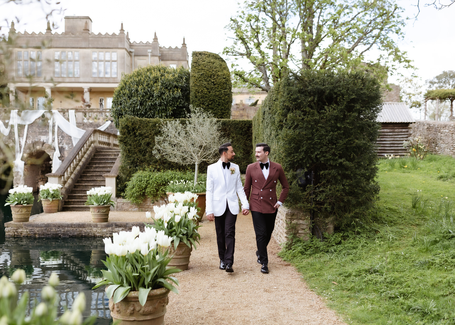 Two grooms in tuxedos, one in white and one in brown, hold hands and walk on a garden path lined with white tulips, with a grand historic building behind—captured perfectly by a country-house wedding photographer.