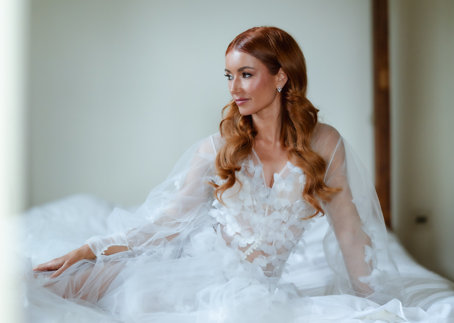 A woman with long, wavy red hair in a sheer, white floral gown sits on a white bed, gazing to the side with a soft expression—captured in the gentle light by a country-house wedding photographer.