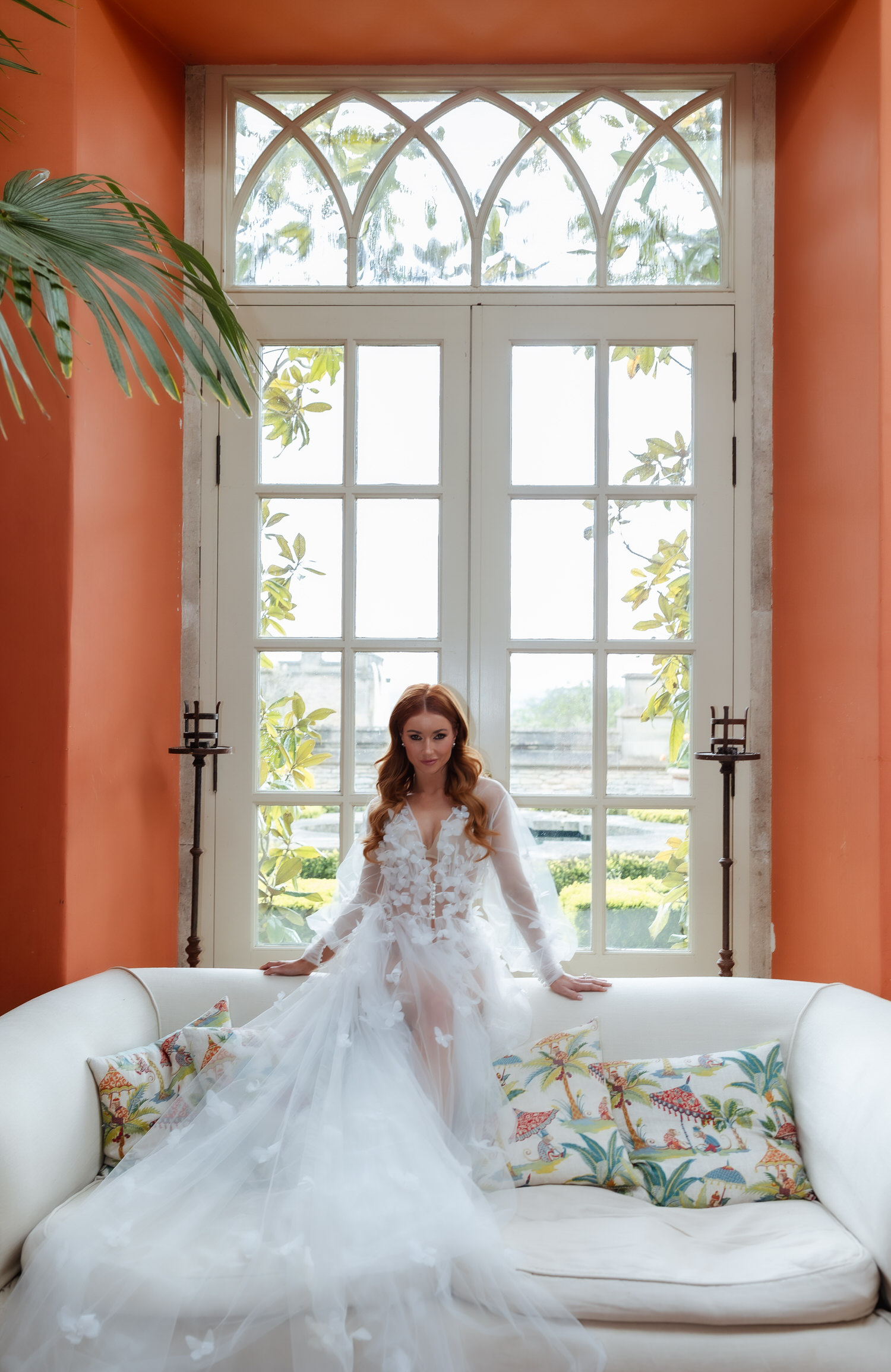 A woman in a flowing white gown sits on a white couch with colorful floral pillows, framed by leafy plants outside the window—an ideal scene for a country-house wedding photographer capturing moments in a bright room with orange walls.
