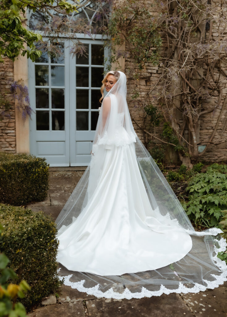 A bride in a white dress and long lace veil stands outdoors on a stone path, looking over her shoulder and smiling. She is surrounded by greenery, with a stone wall and pale blue double doors behind her.