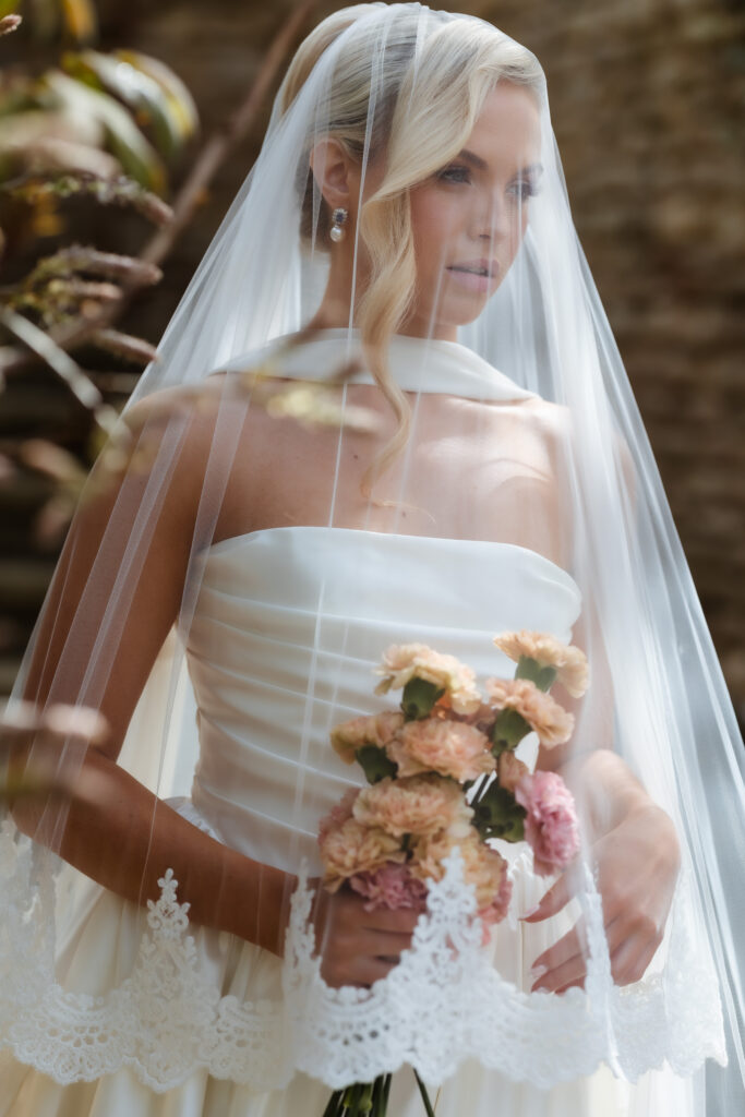 A bride in a strapless white dress and long, lace-edged veil holds a bouquet of pastel pink and peach flowers, standing outdoors with sunlight filtering through trees.