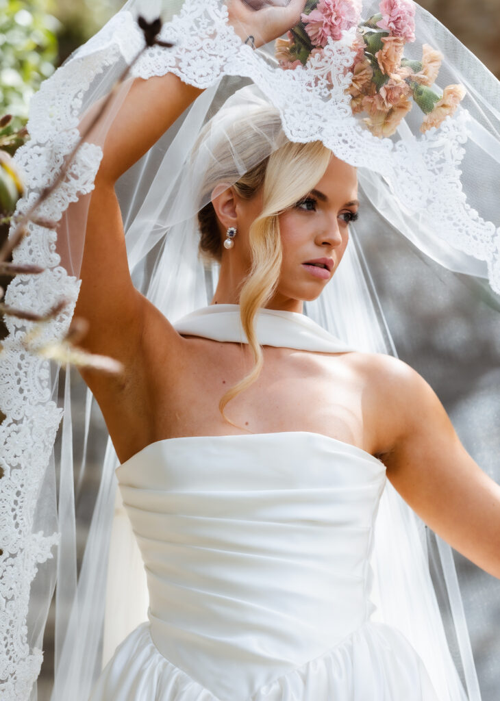 A bride in a white satin dress and lace veil holds up her veil with one hand. She has blonde hair styled with a loose curl and wears pearl earrings, with a bouquet of pink flowers above her head.