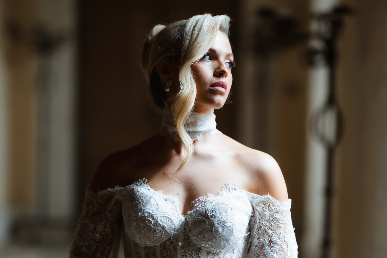 A woman in an off-shoulder lace wedding dress looks thoughtfully to the side. Captured by a Hampshire wedding photographer, she has blonde hair styled in soft waves and pearl earrings, standing in soft, warm indoor lighting.