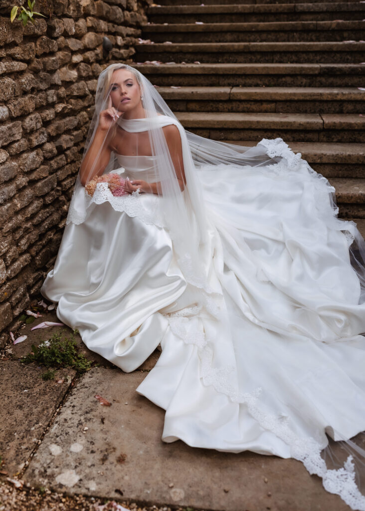 A bride in a long, flowing white wedding dress and veil sits on stone steps, holding a bouquet of pink roses. She gazes thoughtfully upward, with petals scattered around her on the ground.