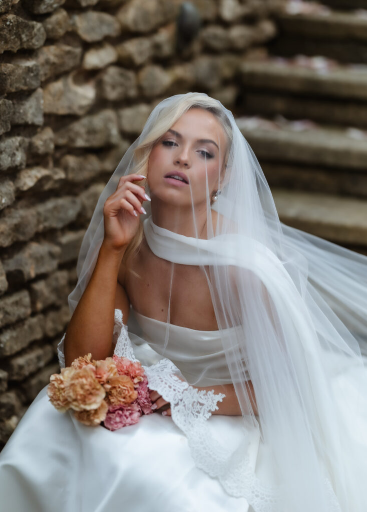 A bride in a white dress and veil sits on stone steps, holding a bouquet of pink and peach flowers. She gazes softly at the camera with one hand near her face.