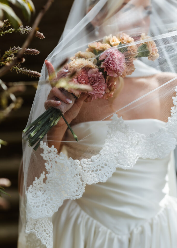 A bride in a white dress and lace veil holds a small bouquet of pink carnations close to her face, partially obscured by the veil.