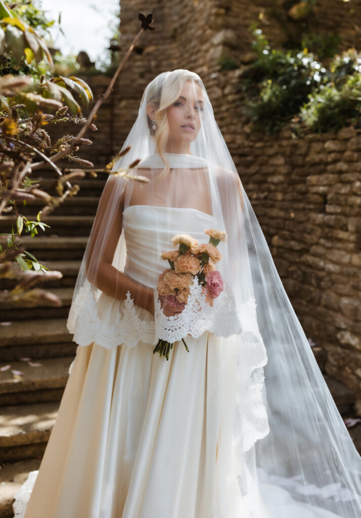 A bride in a white strapless wedding dress and long lace veil holds a bouquet of pink and peach flowers, standing outdoors near stone steps and greenery.