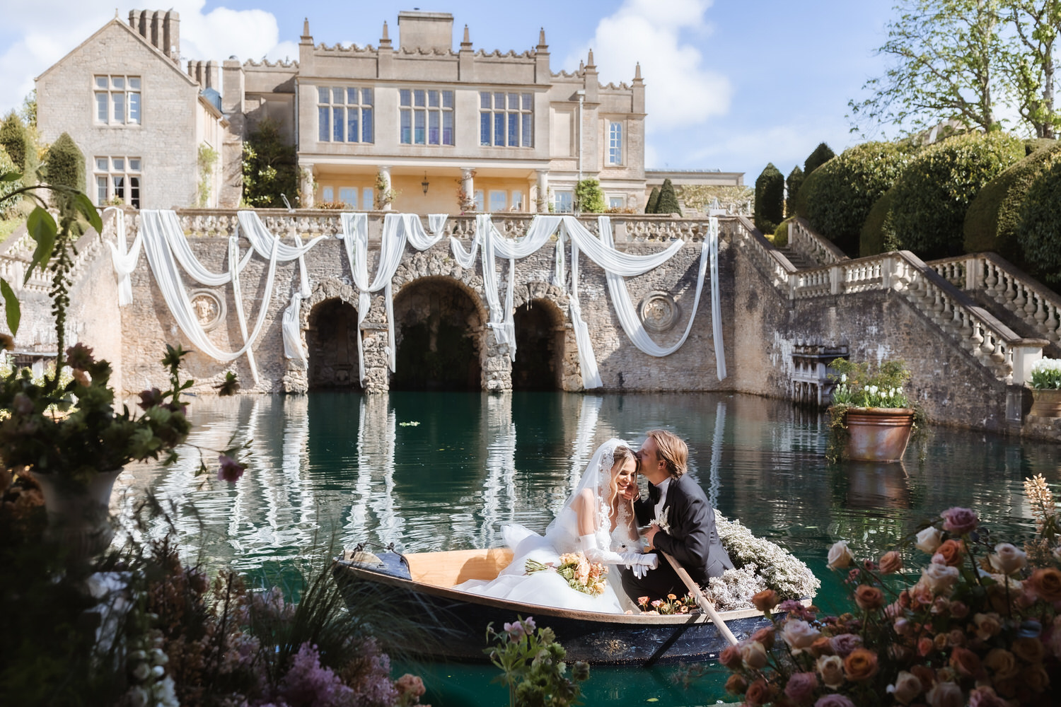 A bride and groom sit in a decorated rowboat on a pond, sharing a joyful moment. Captured by a Hampshire wedding photographer, the scene features a grand manor with white drapery, stone arches, and lush gardens filled with flowers.