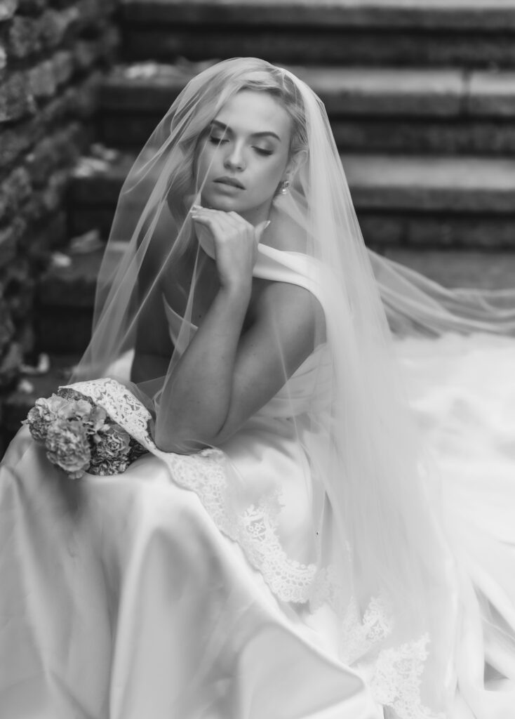 A bride in a wedding dress and veil sits thoughtfully on stone steps, holding a small bouquet of flowers, with her eyes closed and hand resting near her chin. The photo is in black and white.