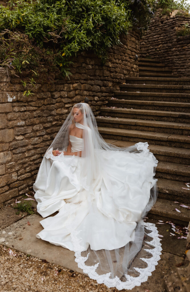 A bride in a white gown with a long veil sits on stone steps surrounded by rustic stone walls and greenery, looking serene and elegant.