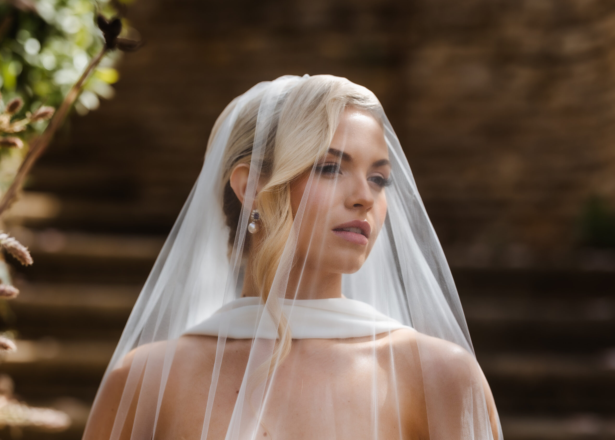 A bride with blonde hair in an elegant updo wears a white veil and pearl earrings, looking slightly to the side against a blurred outdoor background with stone steps.