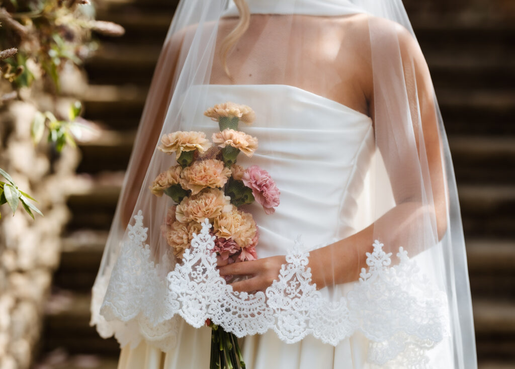 A bride in a white dress and lace-edged veil holds a bouquet of pale pink and peach flowers, standing outdoors with blurred stone steps in the background.