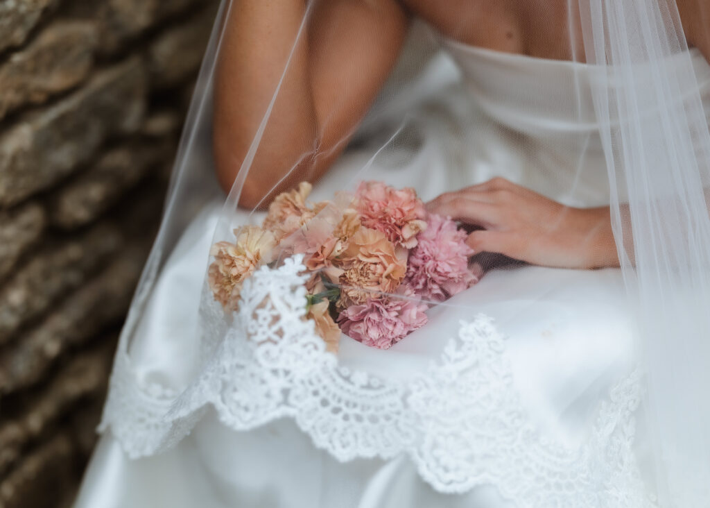 A bride in a white lace-trimmed dress holds a small bouquet of pink and peach flowers, with her veil draped over her arm, sitting next to a stone wall.