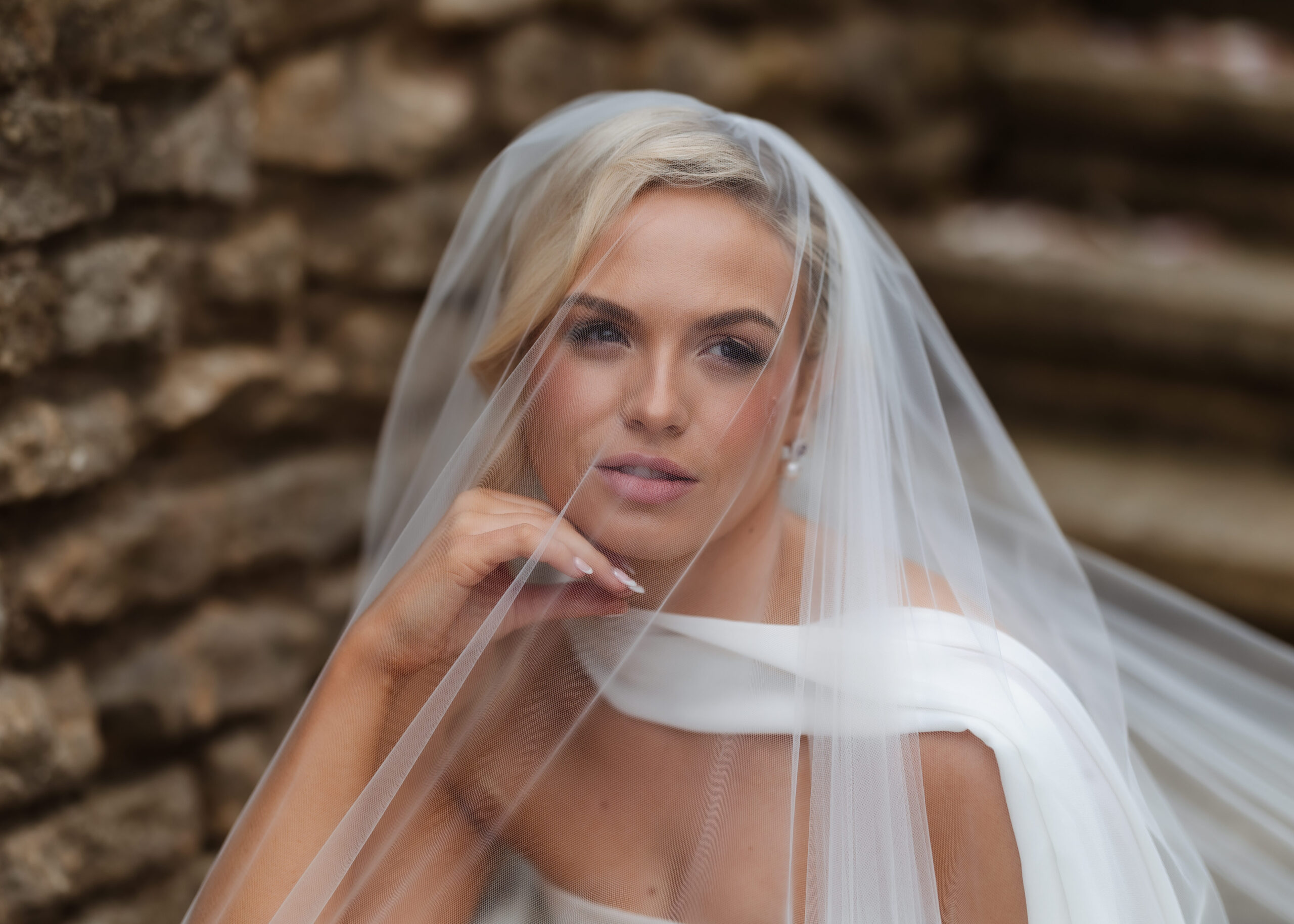 A bride with blonde hair poses thoughtfully, resting her chin on her hand. She wears a white dress and a sheer veil draped over her face, sitting beside a rustic stone wall.
