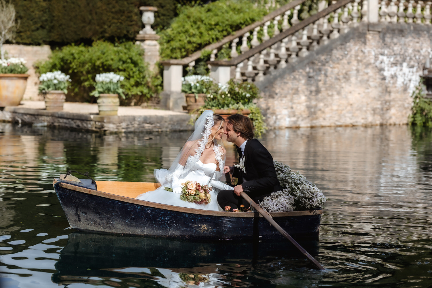 A bride and groom in wedding attire share a kiss while sitting in a flower-adorned rowboat on a calm pond, with stone stairs and lush greenery behind them—captured beautifully by a Hampshire wedding photographer.