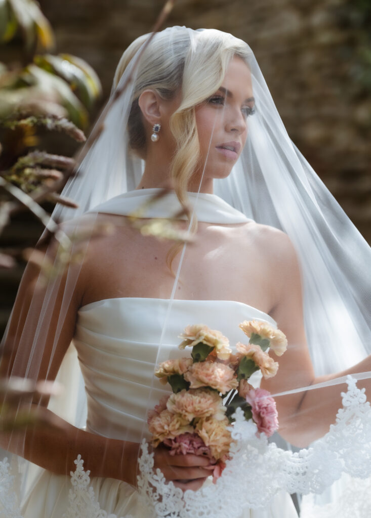 A bride in a white strapless gown holds pastel flowers and gazes thoughtfully, draped in a sheer veil with lace trim, standing outdoors with blurred greenery in the background.