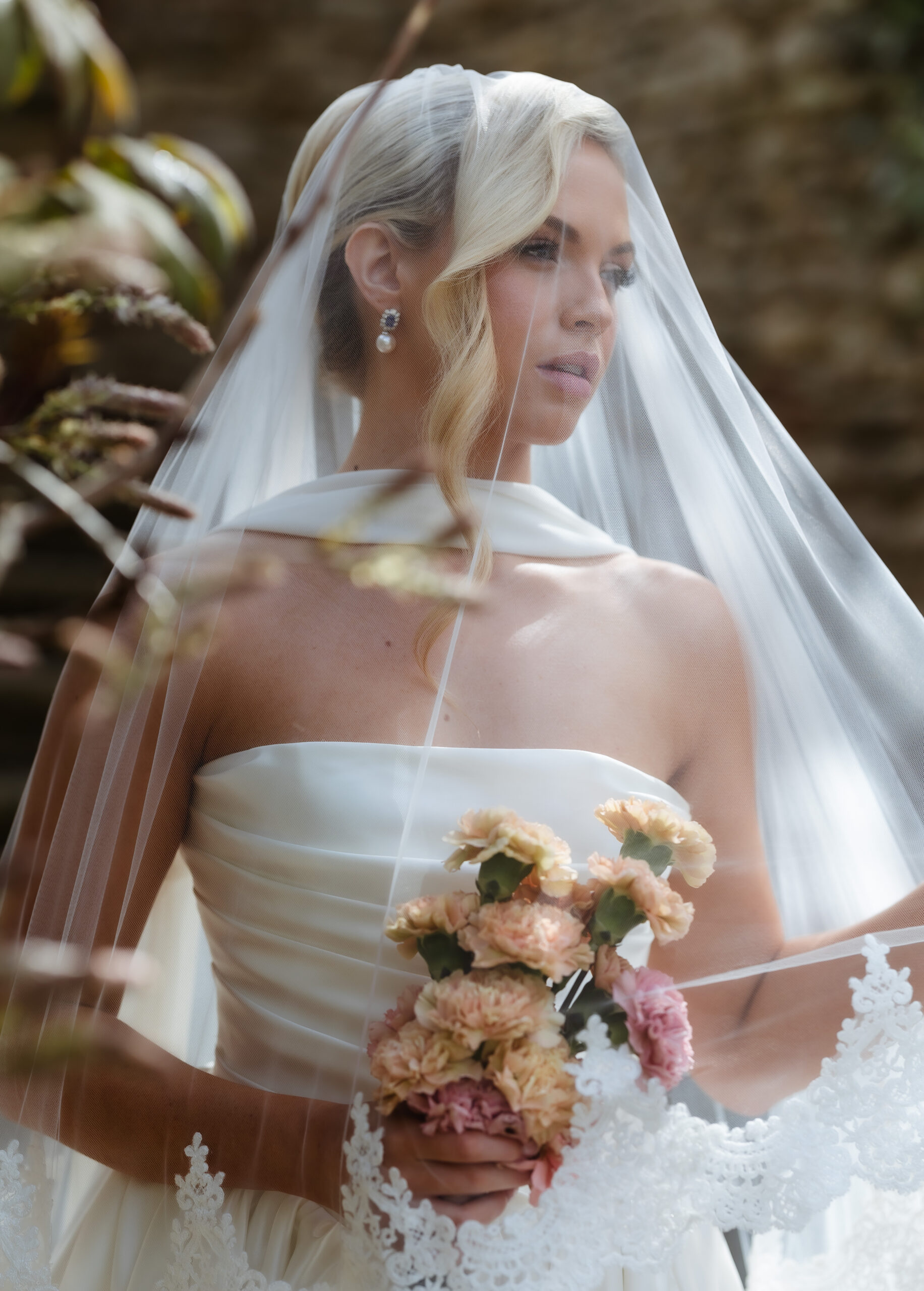 A bride in a white strapless gown holds pastel flowers and gazes thoughtfully, draped in a sheer veil with lace trim, standing outdoors with blurred greenery in the background.