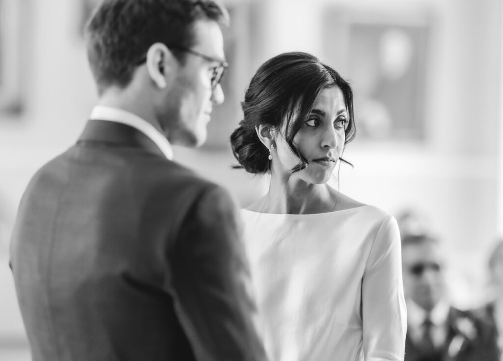 A bride in a white dress and a groom in a suit stand facing each other during a wedding ceremony. The bride looks thoughtfully to the side. The image is in black and white.
