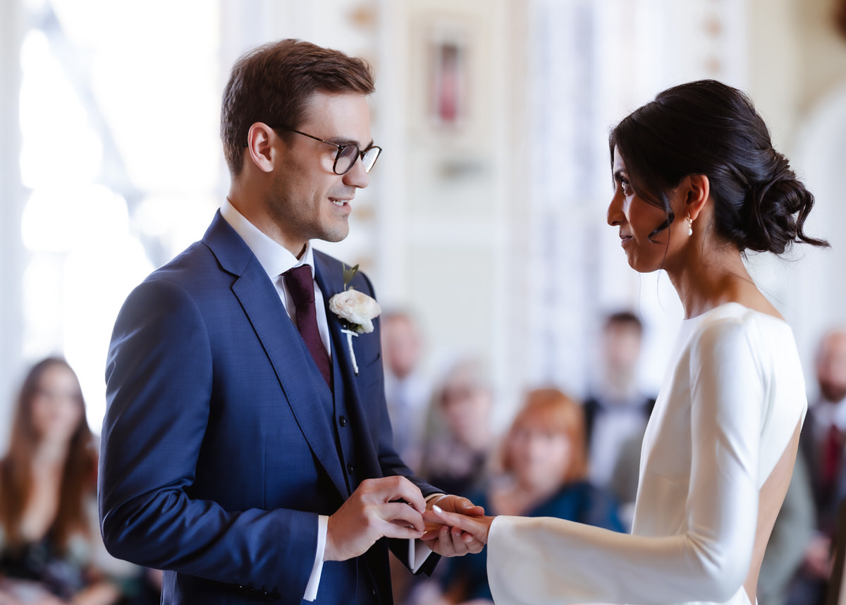 A groom in a blue suit places a ring on the bride’s finger during a wedding ceremony. The bride wears a white dress, and they look at each other smiling. Guests are blurred in the background.