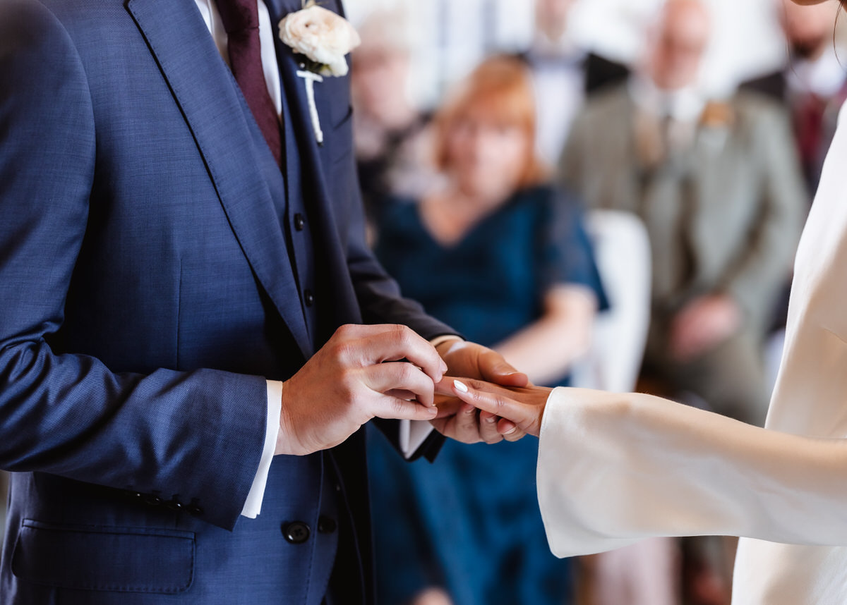 A person in a blue suit places a ring on another persons finger during a wedding ceremony, with a small group of blurred guests seated in the background.