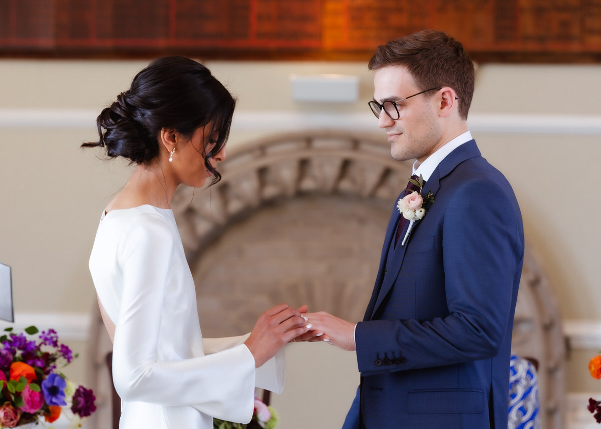 A bride in a white dress places a ring on the grooms finger during an indoor wedding ceremony. They face each other, surrounded by colorful flowers, with a soft, warm background.