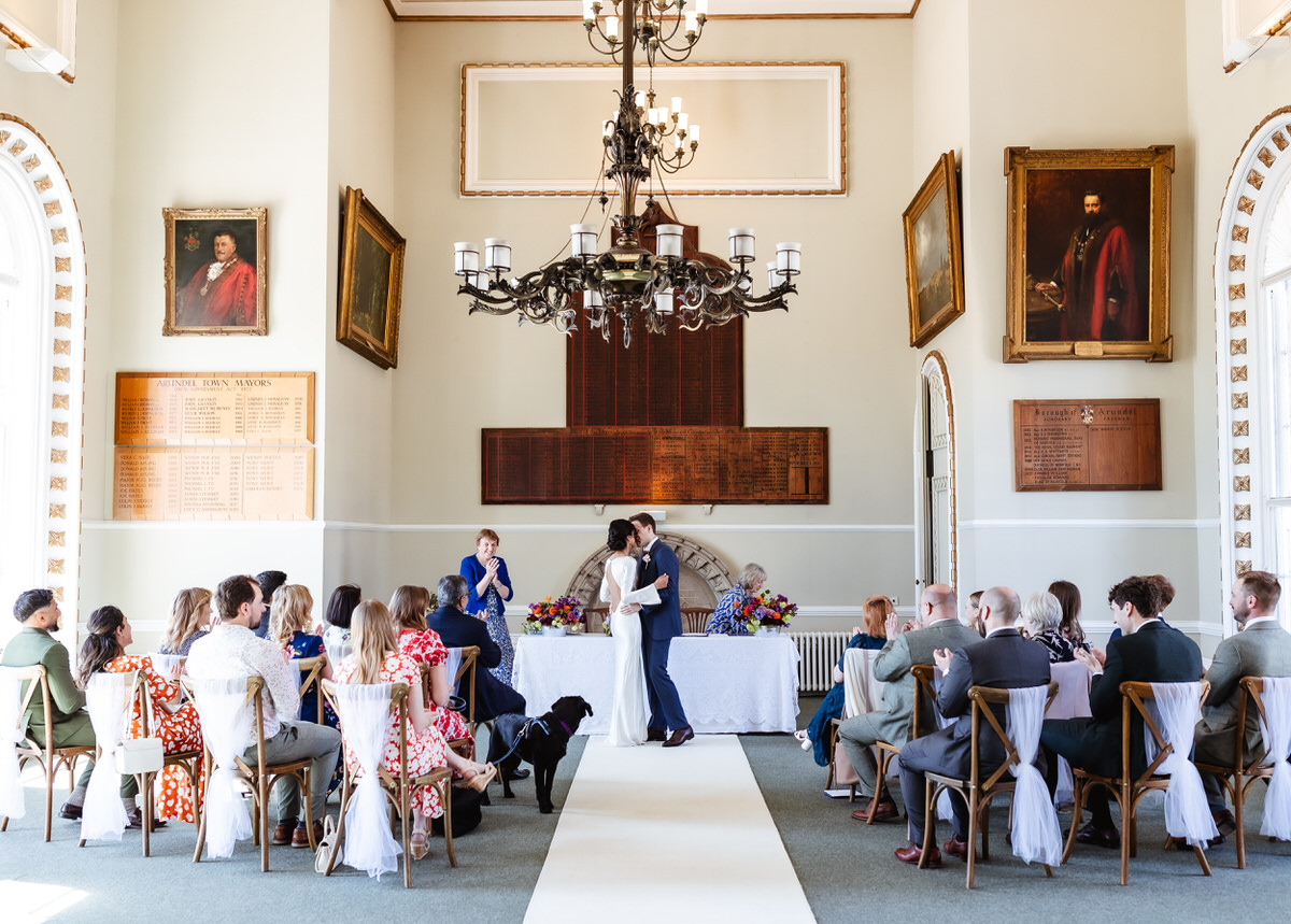 A couple shares a kiss at the front of a bright ceremony room as guests seated on both sides watch. A black dog lies in the aisle. Large portraits and chandeliers decorate the high-ceilinged space.