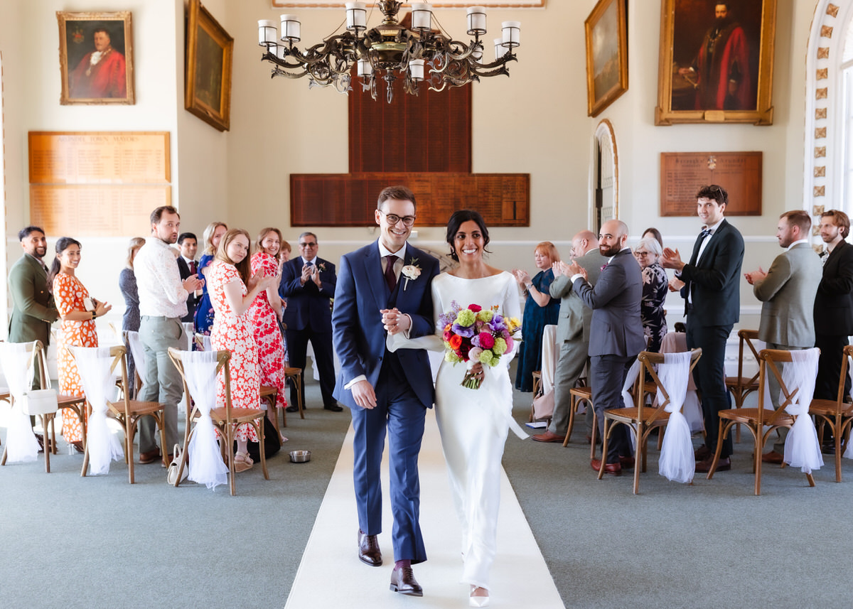 A newly married couple walks arm in arm down the aisle, smiling, as guests in formal attire stand and applaud in a bright, elegant room decorated with portraits and chandeliers.
