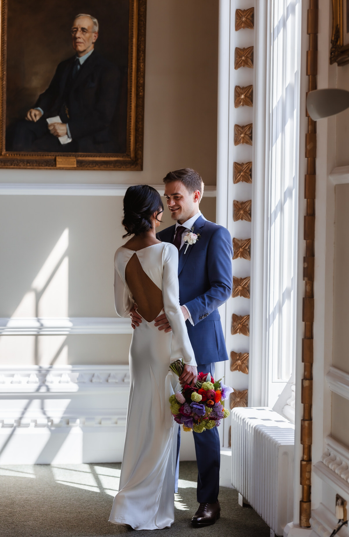 A bride in a long white dress and groom in a blue suit stand close together by a sunlit window, smiling at each other. The bride holds a colorful bouquet. A portrait painting hangs on the wall behind them.