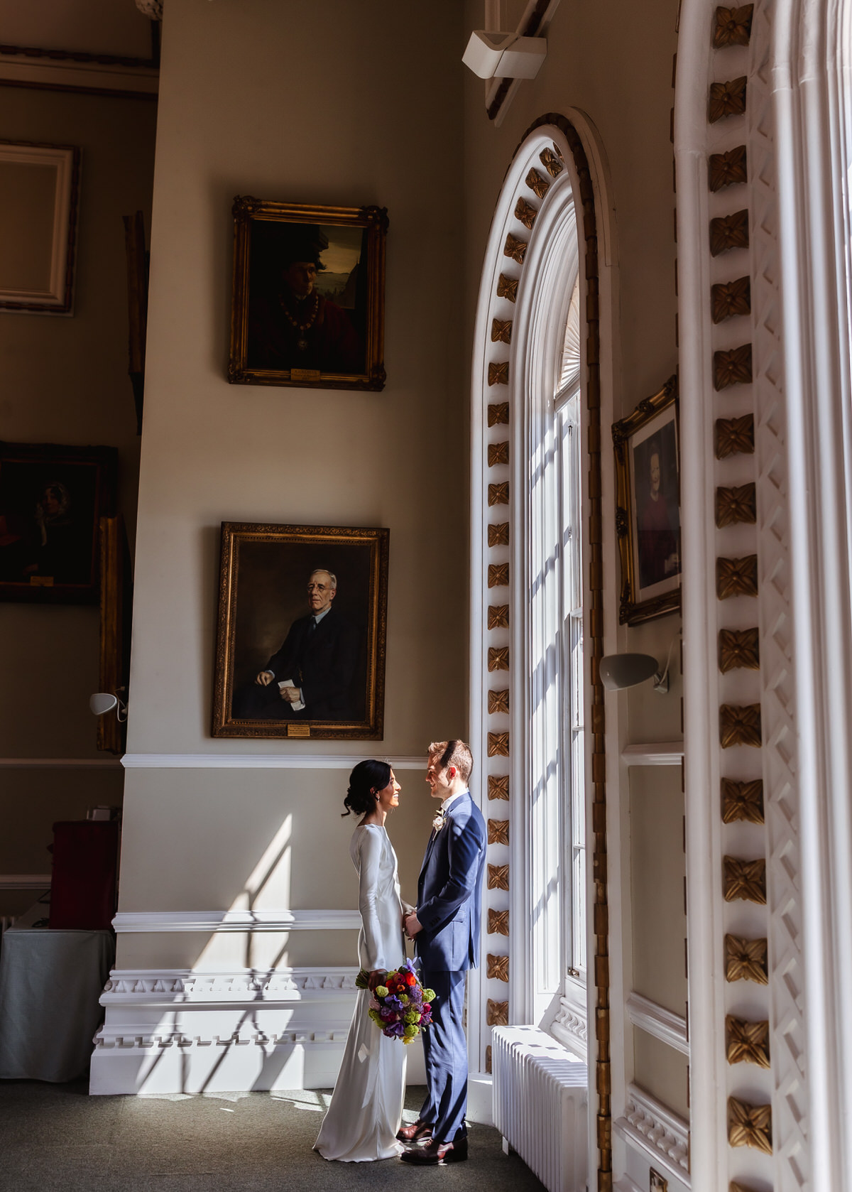 A bride and groom stand facing each other by a large arched window, bathed in sunlight, inside an elegant room decorated with classical portraits on the walls. The bride holds a colorful bouquet.