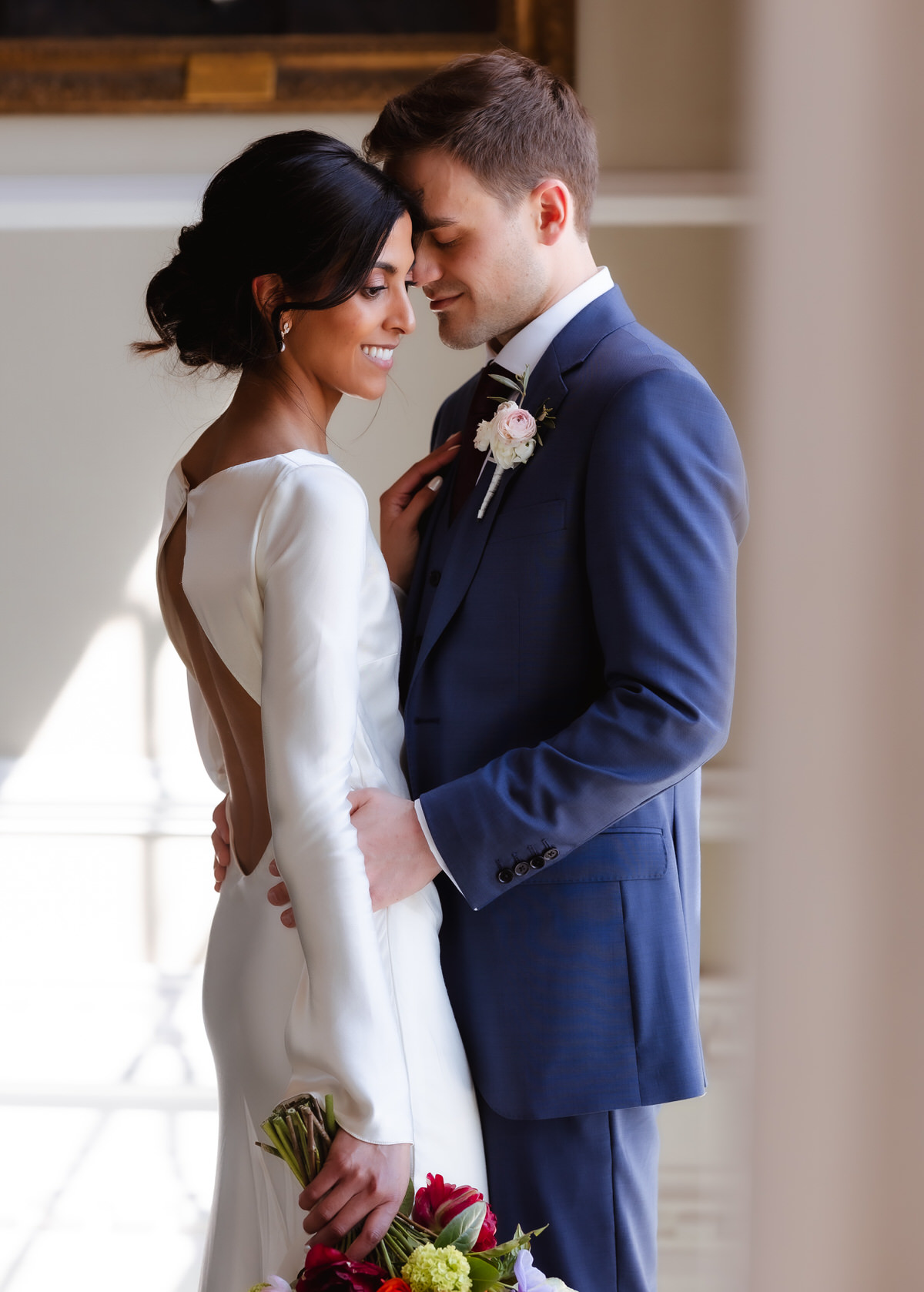 A bride in a white dress holds a bouquet as she stands close to a groom in a blue suit. They smile gently, foreheads touching, in a softly lit, elegant indoor setting.