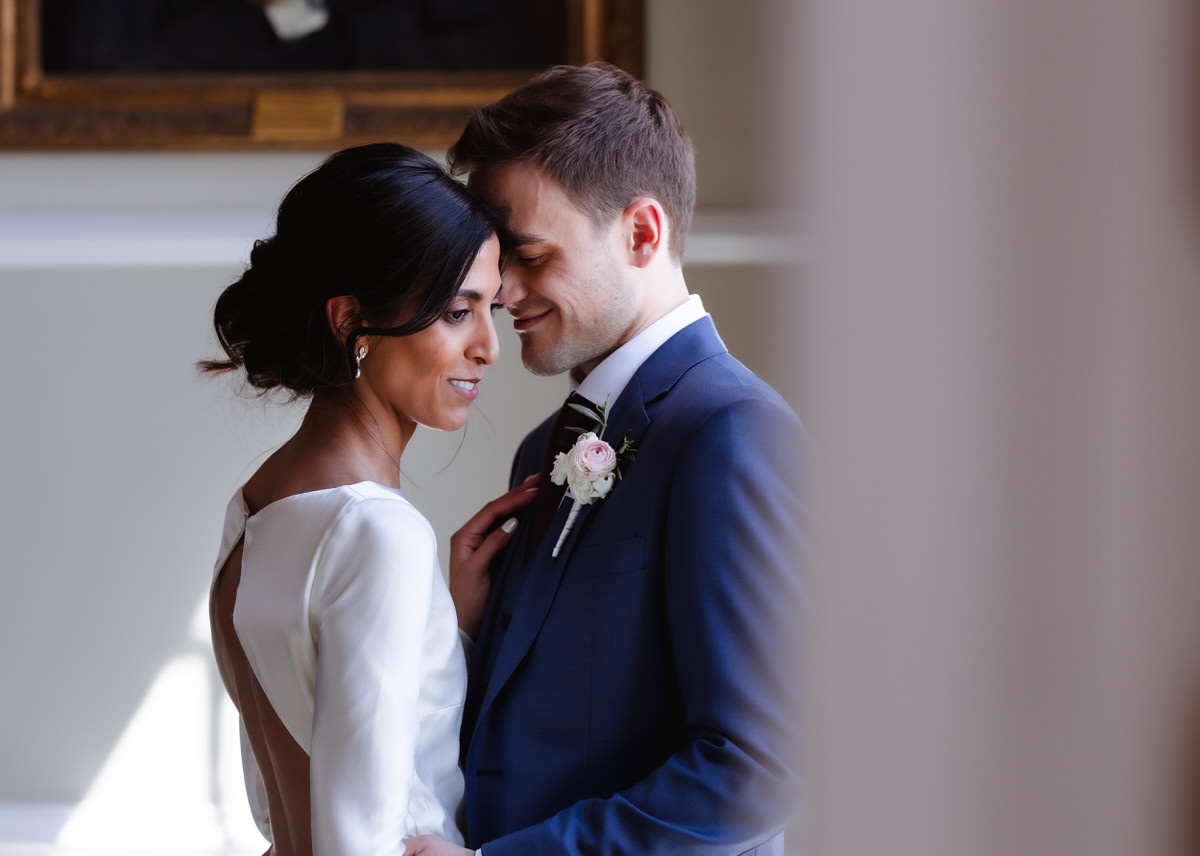A bride in a white dress and a groom in a blue suit share an intimate, joyful moment indoors, smiling and gently touching foreheads, with soft natural light illuminating them.