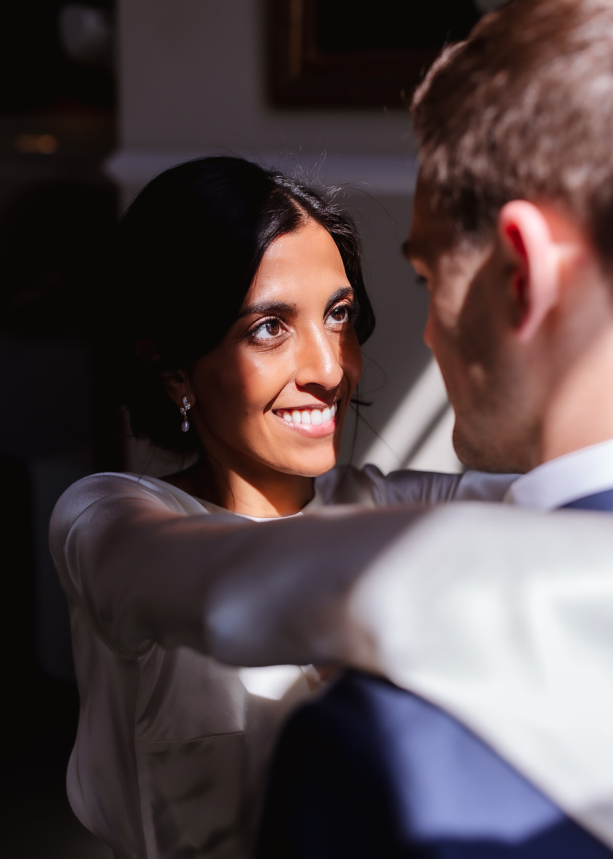A bride in a white dress smiles warmly while holding her partner, who is wearing a dark suit, during a close and intimate moment, with sunlight softly illuminating her face.