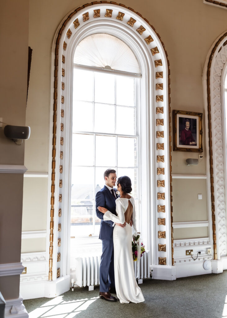 A couple dressed in wedding attire embraces in front of a tall, arched window with gold trim, sunlight streaming in. The bride holds a bouquet and the groom wears a suit. A framed portrait hangs on the wall beside them.