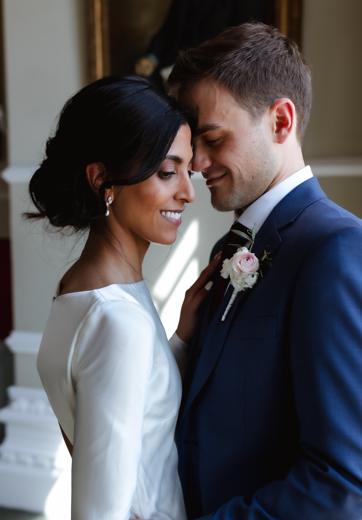A bride and groom stand close together, smiling with their foreheads touching. The bride wears a white dress and pearl earrings; the groom wears a blue suit with a boutonniere. Soft natural light fills the scene.