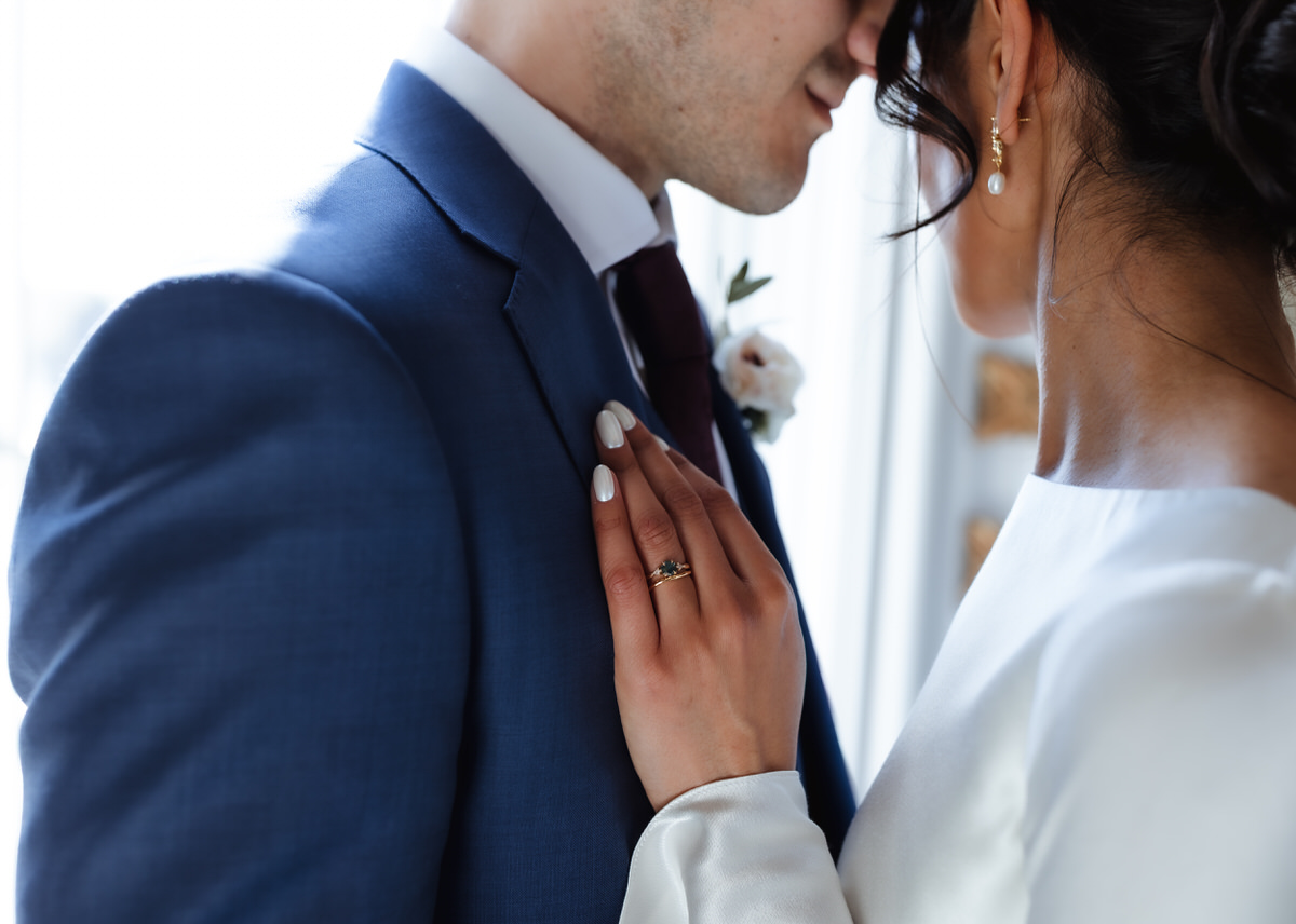 A close-up of a bride and groom embracing, with the bride’s hand resting on the groom’s chest, showing her wedding ring. The groom wears a blue suit and boutonniere; both appear to be smiling softly.