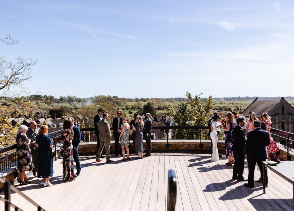 A group of people dressed in formal attire stand and mingle on a large outdoor terrace with a scenic countryside view under a clear, sunny sky.