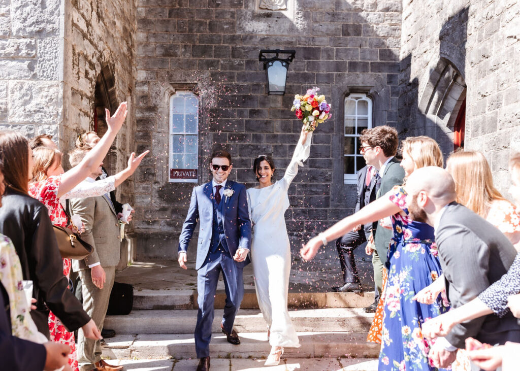 A smiling bride and groom walk hand in hand outside a stone building, surrounded by guests tossing confetti. The bride raises her bouquet triumphantly, and sunlight brightens the joyful scene.