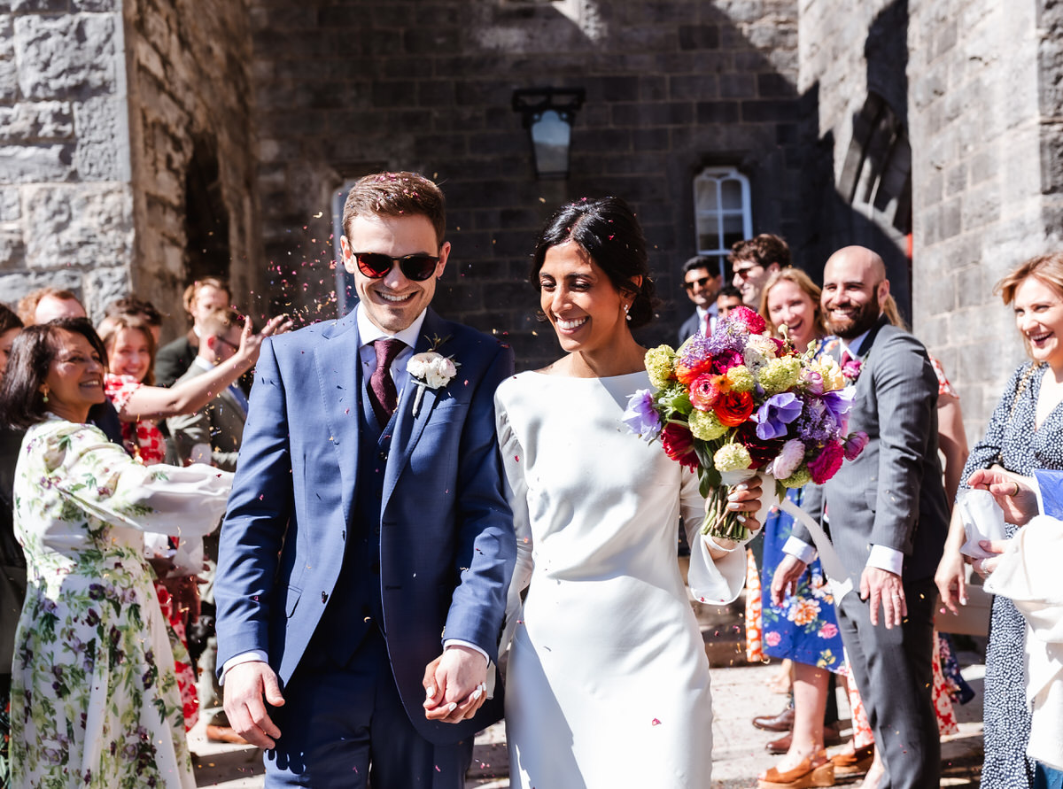 A bride and groom, smiling and holding hands, walk outside a stone building as guests cheer and throw confetti. The bride holds a bright bouquet of flowers; both are dressed formally and appear joyful.