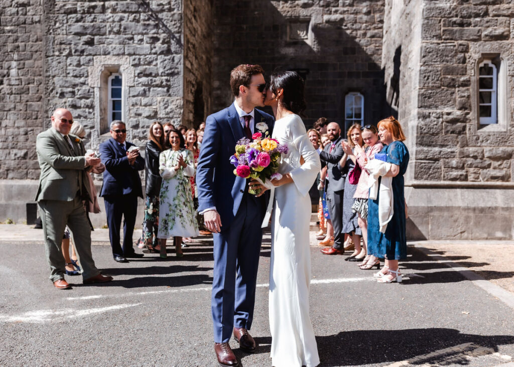 A bride and groom share a kiss outside a stone building, holding a colorful bouquet, while wedding guests stand behind them clapping and smiling.