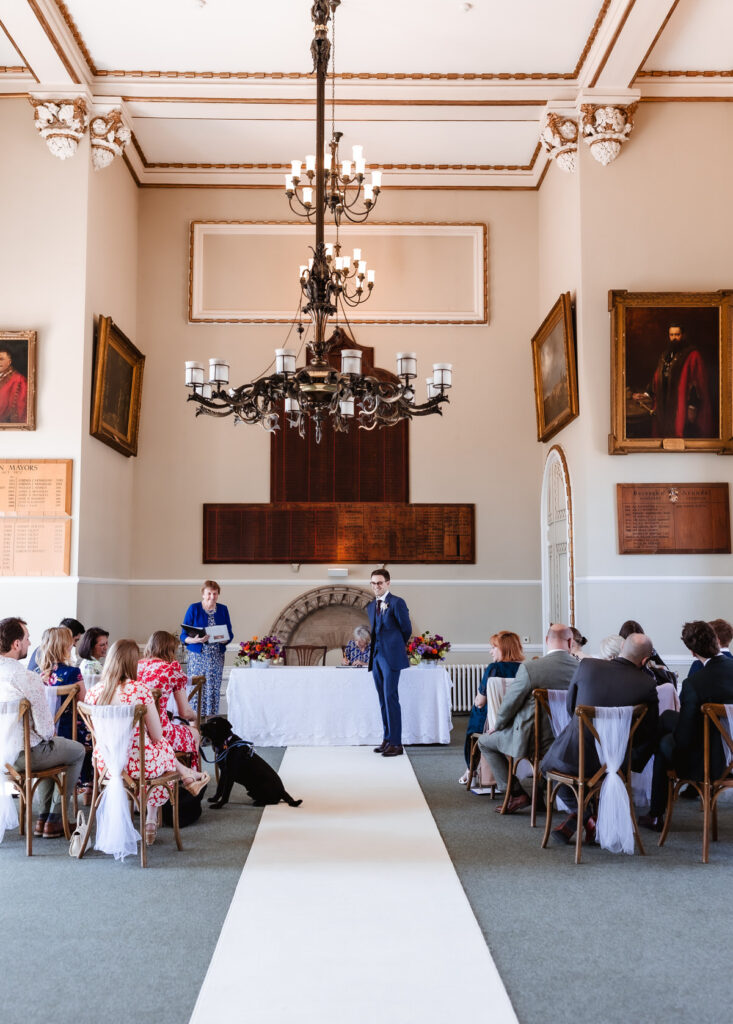 A man in a blue suit stands at the front of a formal room with high ceilings, chandeliers, and portraits on the walls. Guests are seated facing him, and a black dog sits in the aisle on a white runner.