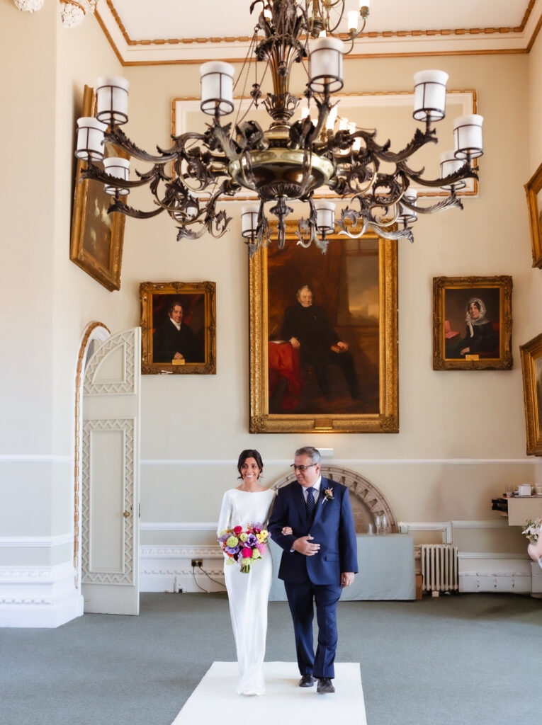A bride in a white dress holding a colorful bouquet walks down the aisle with an older man in a suit inside an elegant room with large portraits and a grand chandelier.