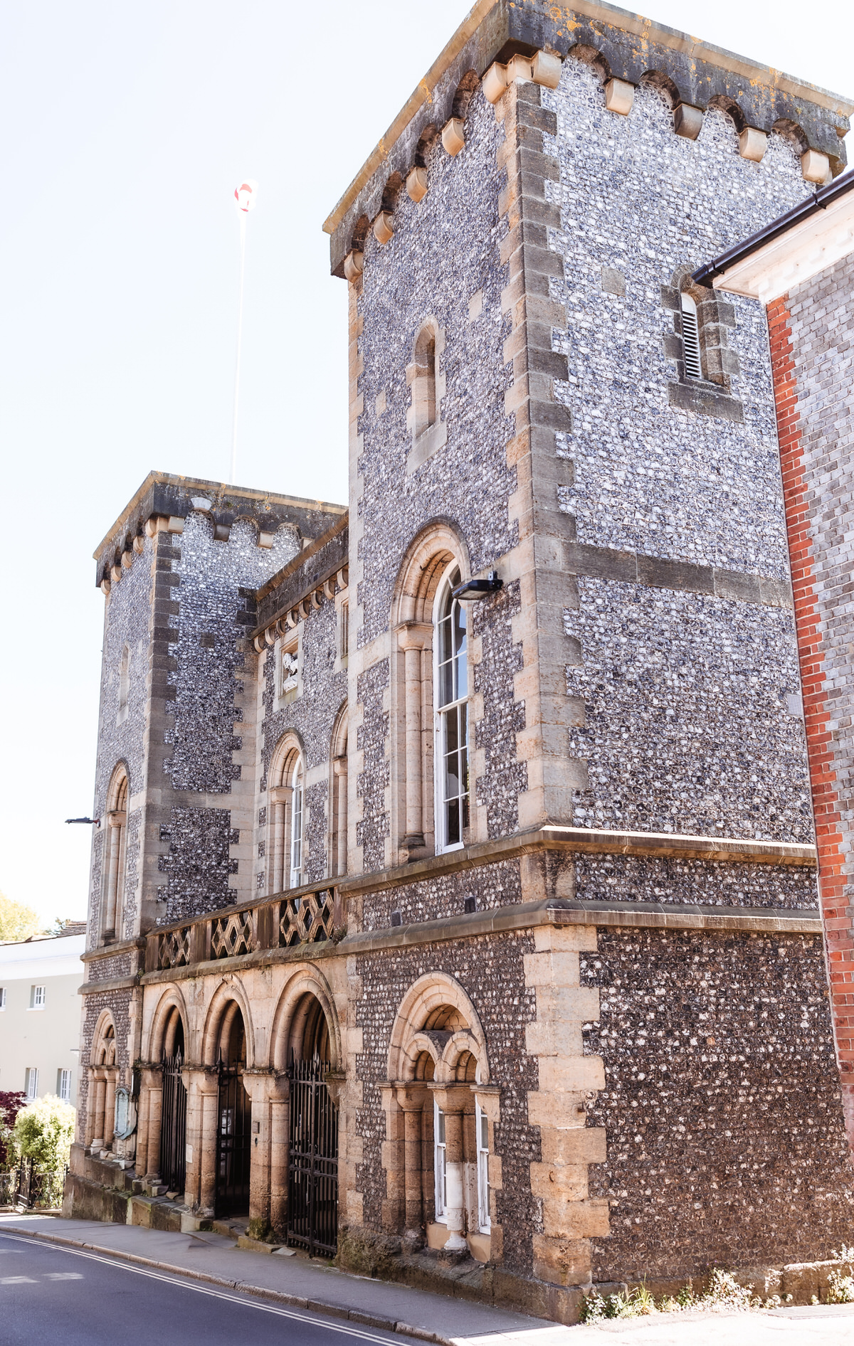 A historic stone building with two tall, square towers featuring arched windows and detailed stonework, set against a clear blue sky. A flagpole extends from the roof of one tower.