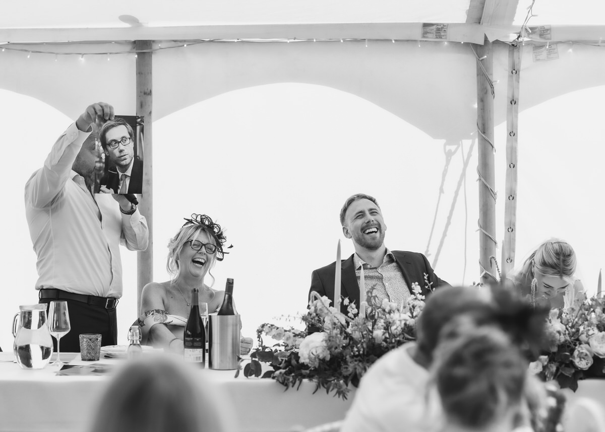 A man holds up a large photo, making guests at a decorated table laugh during a wedding reception in a bright, tented venue.