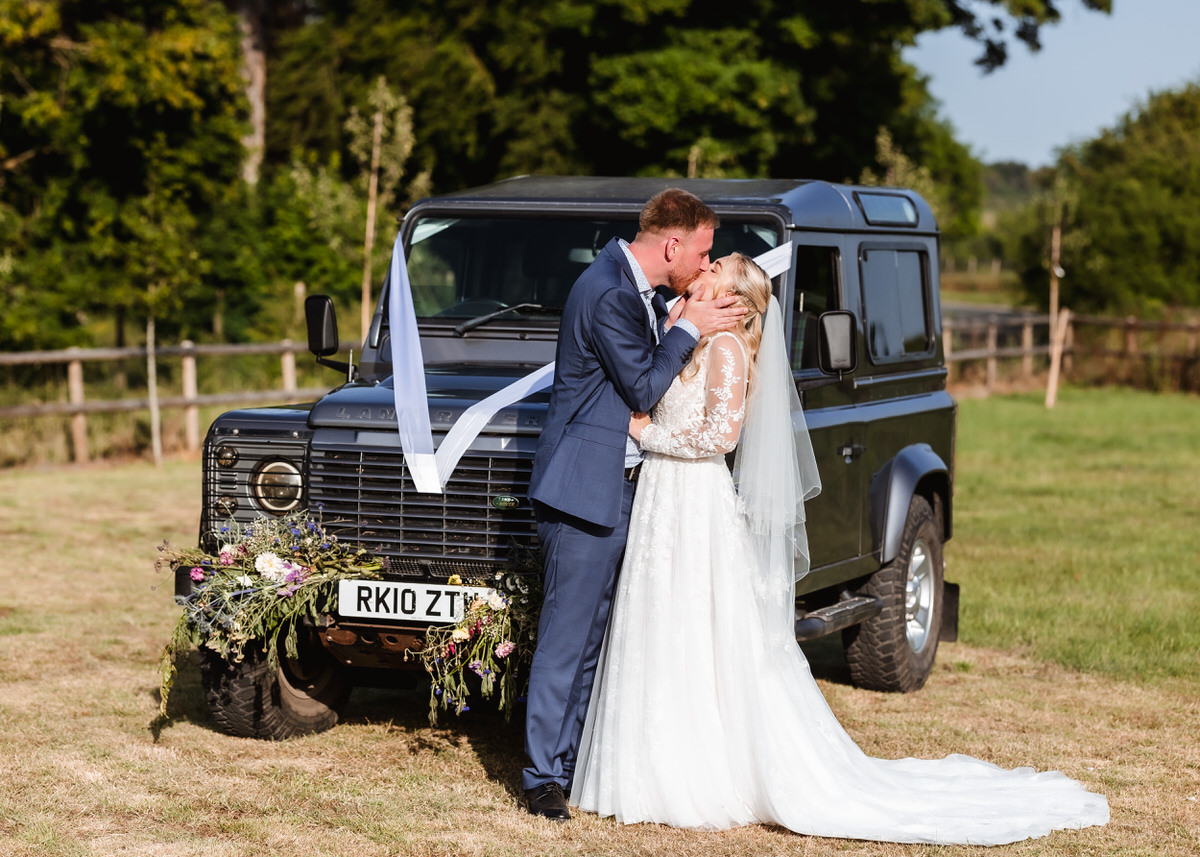 A bride and groom kiss in front of a decorated black SUV in a grassy field. The groom wears a blue suit; the bride wears a white gown and veil. Trees and a wooden fence are in the background.